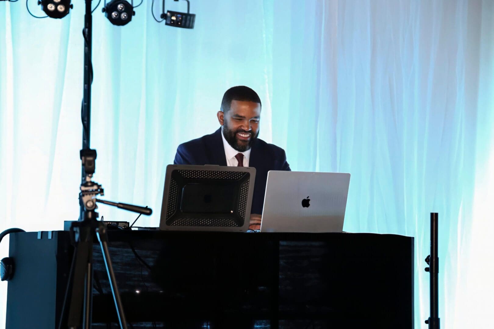 Music disc jockey smiling, looking at two laptop computers, both placed on a DJ booth, with a light blue curtain behind him.